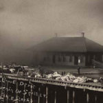 The Great Seattle Fire of June 6, 1889 was a significant turning point in the city’s history and changed both the physical and political landscapes. In this image goods are piled on the dock while spectators watch the fire in progress near the Seattle, Lakeshore & Eastern Railway depot. (MOHAI)